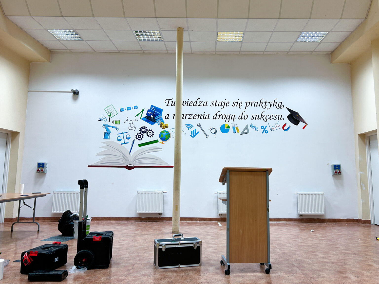 Ścienny druk UV przedstawiający otwartą książkę oraz symbole wiedzy i rozwoju. Tego typu nadruk na ścianie doskonale podkreśla charakter placówek edukacyjnych, bibliotek i sal wykładowych, tworząc przyjazne i kreatywne środowisko