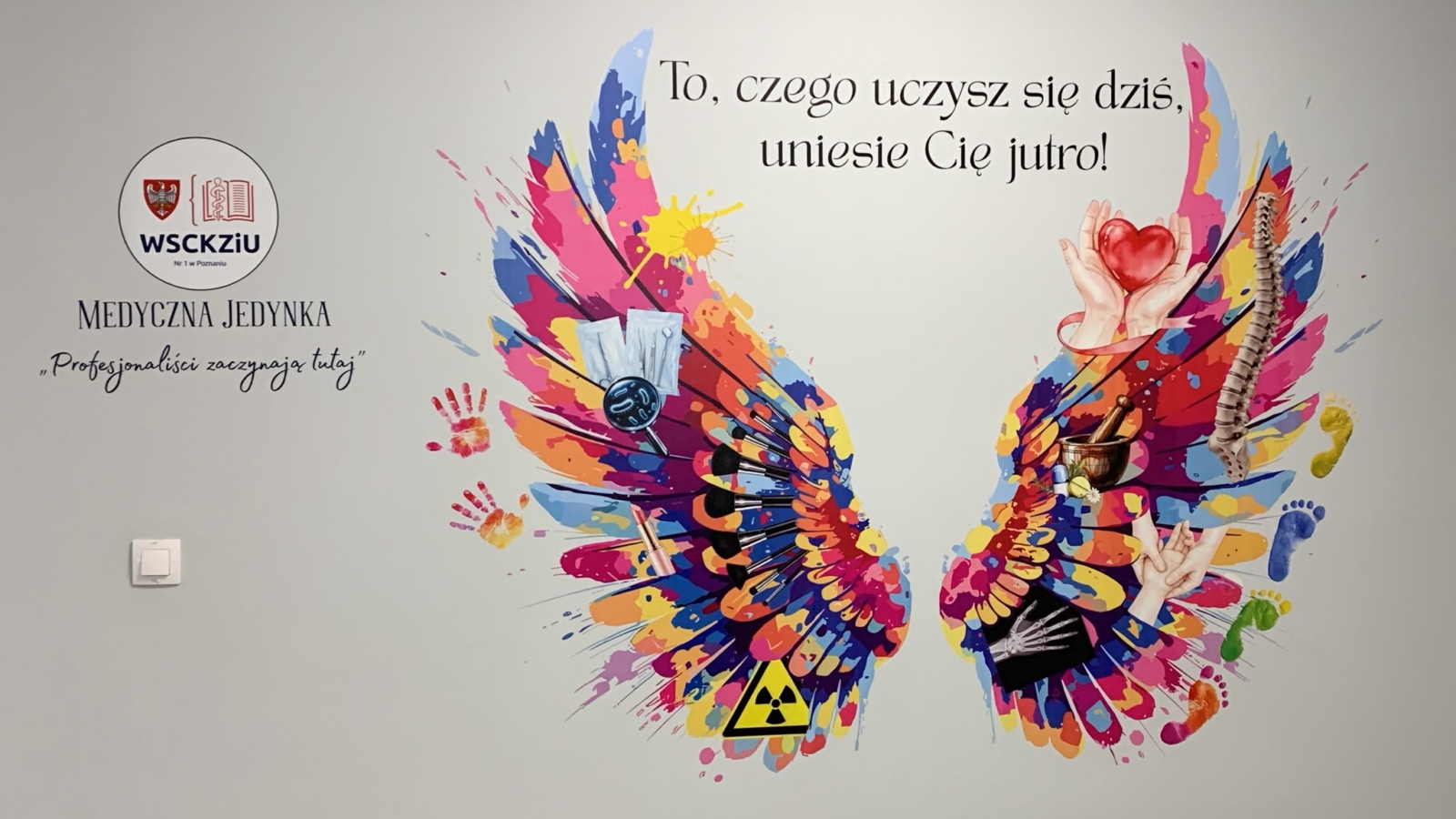 Kolorowe skrzydła wydrukowane bezpośrednio na ścianie dla WSCKZiU Nr 1 w Poznaniu, z symbolami medycznymi i hasłem „To, czego uczysz się dziś, uniesie Cię jutro!”.